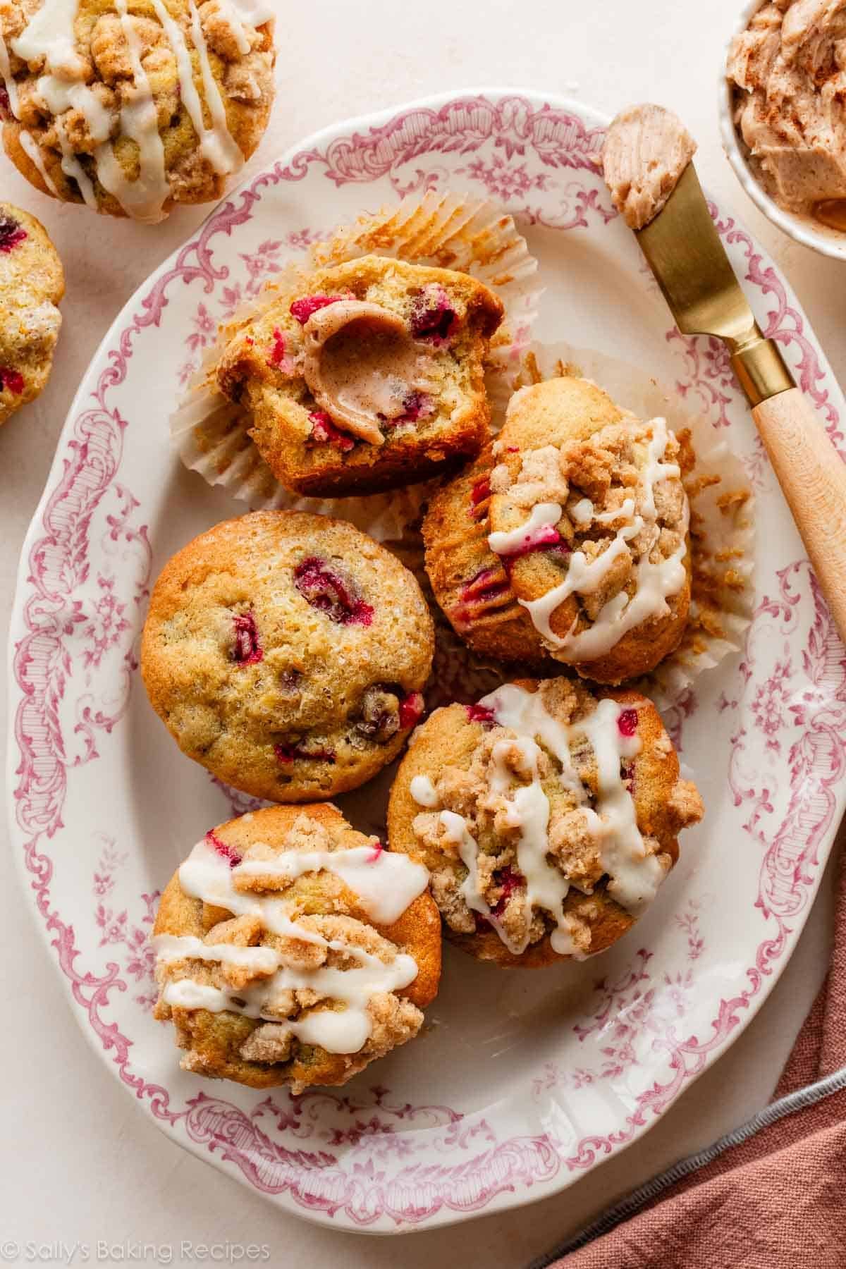 muffins à l'orange et aux canneberges sur une assiette de service florale rose avec du beurre à la cannelle sur un muffin coupé en deux.