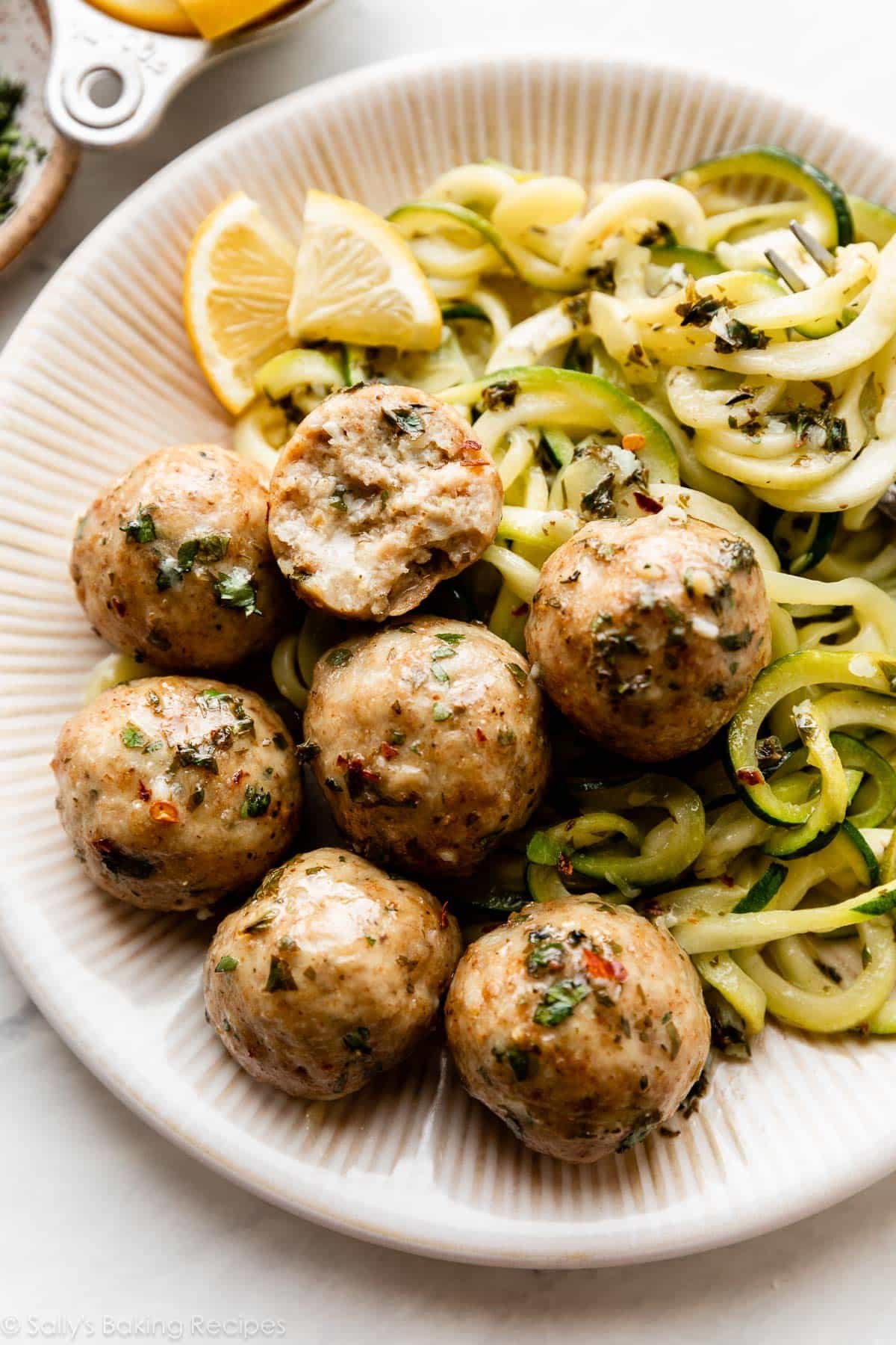 boulettes de poulet avec des herbes sur le dessus sur une assiette avec des nouilles de courgette et des tranches de citron.