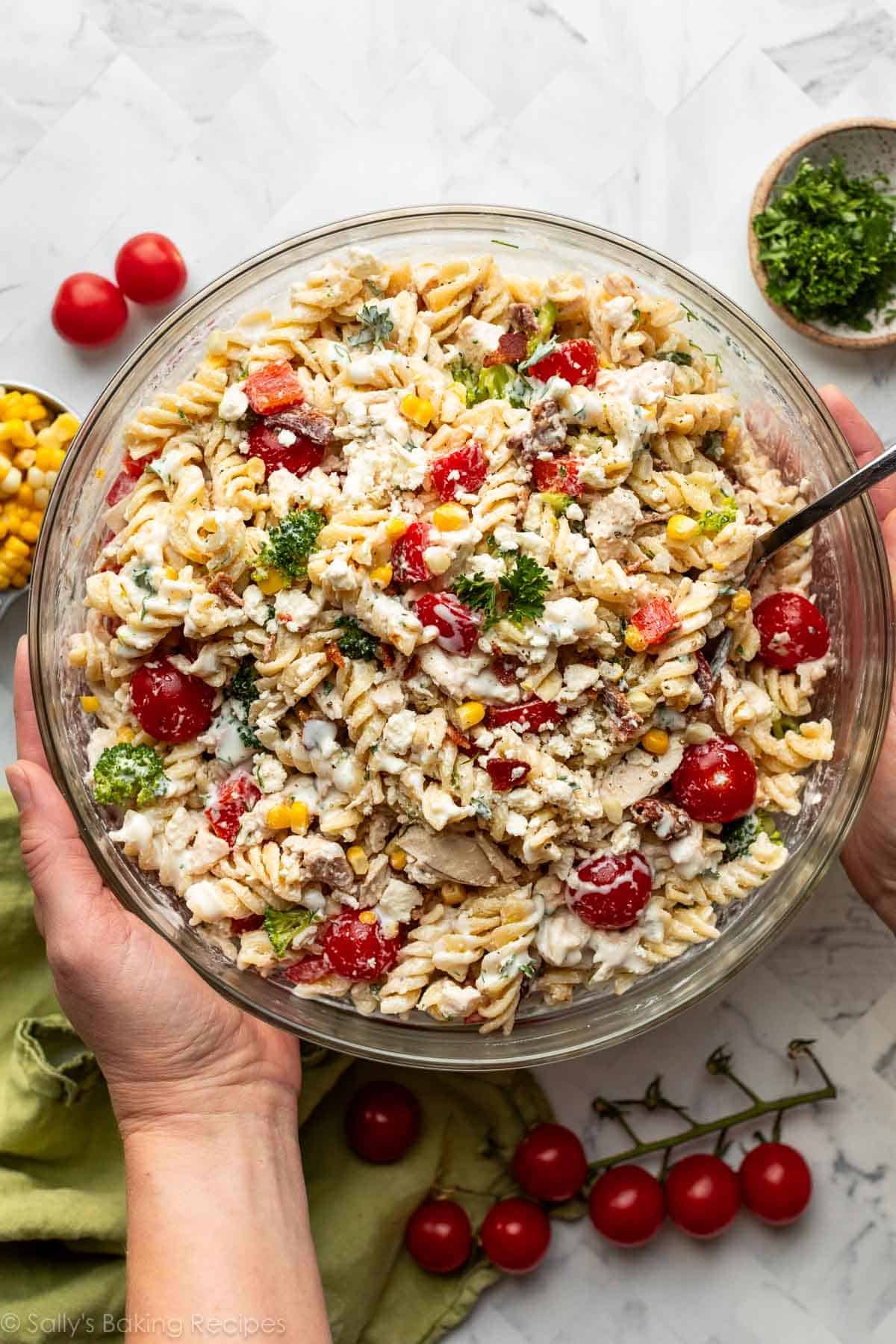 mains prenant un bol en verre de salade de pâtes avec tomates, brocoli, maïs et poivrons rouges