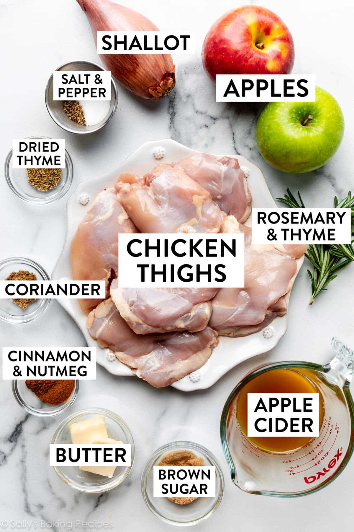 ingrédients sur un comptoir en marbre comprenant des pommes, des cuisses de poulet sur une assiette, du sucre brun, du cidre de pomme, du sel, du poivre et de l'échalote.