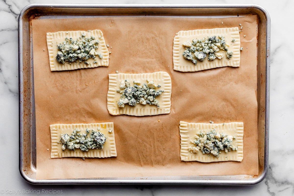 Fromage gorgonzola émietté sur les 5 rectangles de pâte sur une plaque de cuisson tapissée.