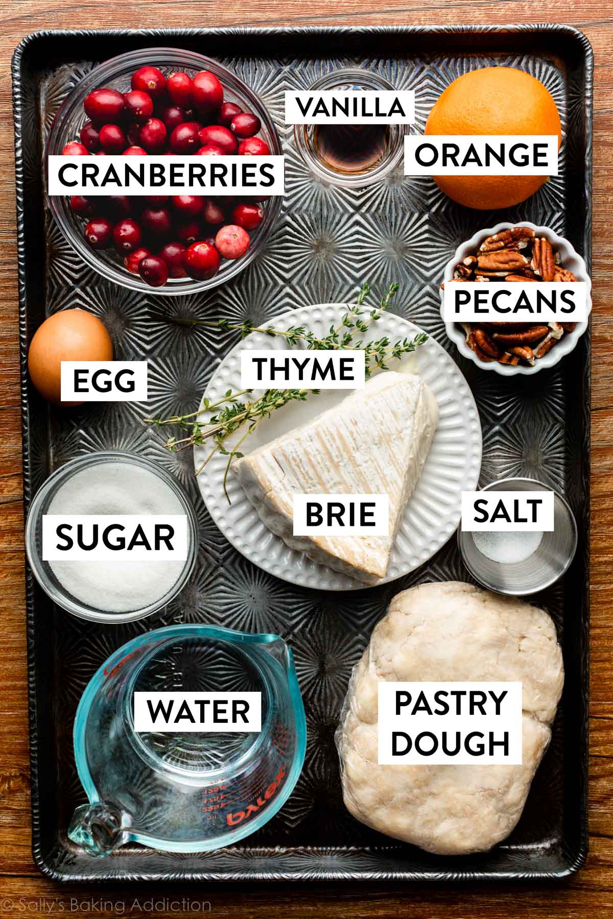 ingrédients sur une plaque de cuisson sombre incluant cranberries, orange, brie, thym, pâte, eau, sucre et pacanes.