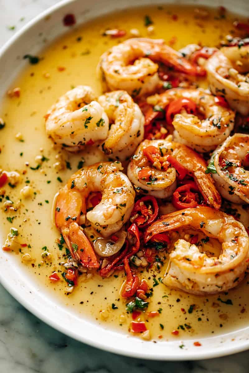 Sur une assiette blanche, une portion de crevettes à l'ail et au beurre épicé dans un lit de sauce avec des morceaux de piment rouge, du persil vert.