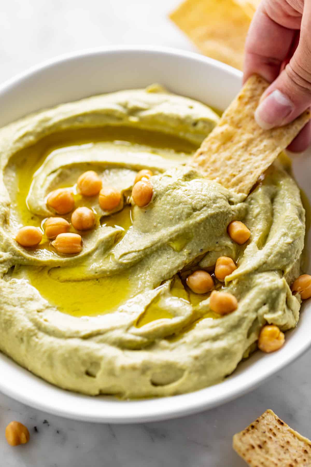 Une chip de maïs trempe dans un bol blanc rempli de Houmous à l'Avocat, arrosé d'huile d'olive et garni de pois chiches entiers.