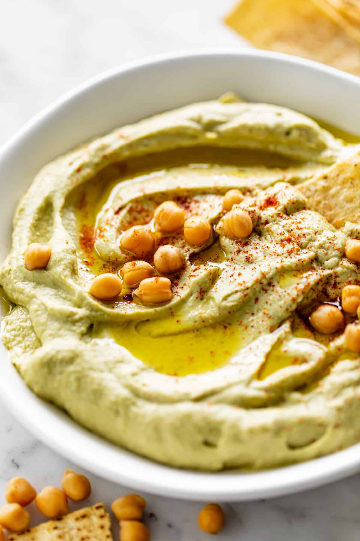 Houmous à l'avocat servi dans un bol blanc, arrosé d'huile d'olive et garni de 16 pois chiches entiers.