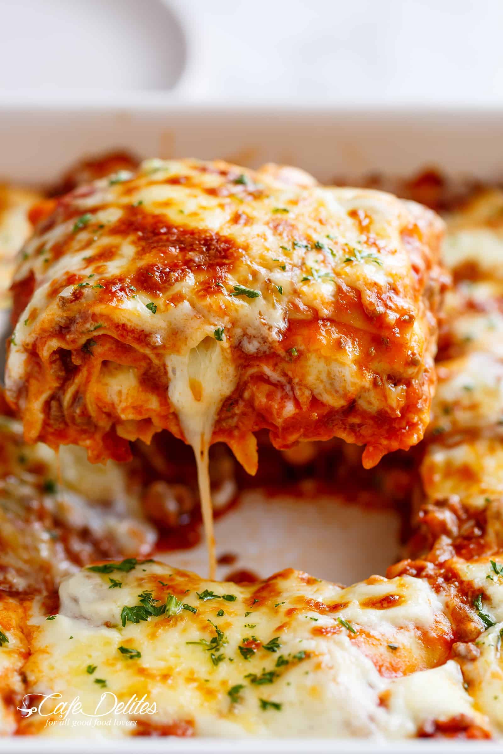 Une portion de lasagne est retirée d'un plat de cuisson blanc, avec un fil de fromage fondu s'étirant. La lasagne est surmontée d'une généreuse couche de fromage mozzarella fondu et saupoudrée de persil haché. Les couches de pâtes, de sauce à la viande et de fromage sont clairement visibles, montrant l'intérieur crémeux et savoureux de la lasagne.