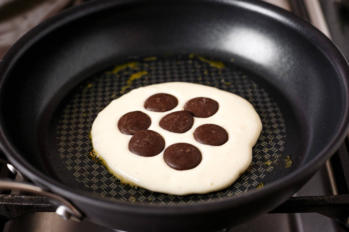 Cuisson de la pâte à crêpes étalée uniformément en forme de crêpe, dans une poêle noire avec des boutons de chocolat placés sur le dessus.