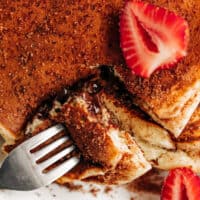 vue de haut d'une pile de crêpes, décorée de sucre à la cannelle et de tranches de fraises fraîches, une fourchette ouvrant un bord coupé de la pile pour voir le centre de chocolat coulant.