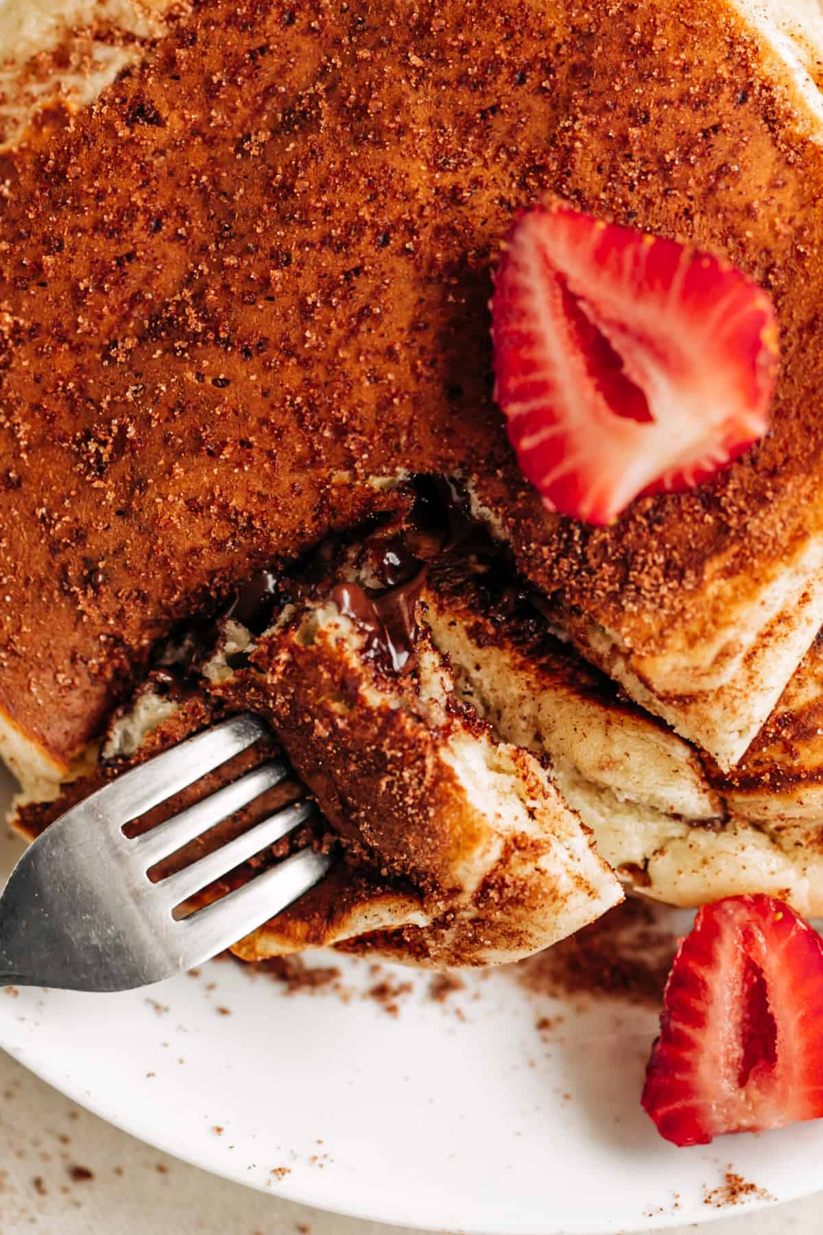 vue de haut d'une pile de crêpes, décorée de sucre à la cannelle et de tranches de fraises fraîches, une fourchette ouvrant un bord coupé de la pile pour voir le centre de chocolat coulant.