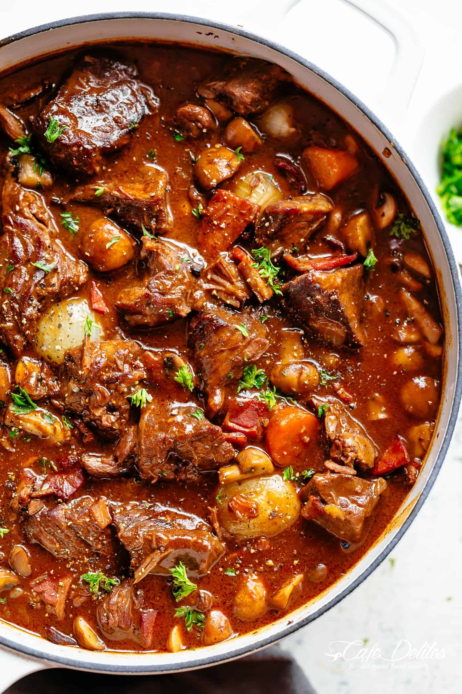 Des morceaux de boeuf tendres mijotés dans un plat en fonte blanc contre un fond blanc. Mijotés dans une sauce riche au vin rouge.