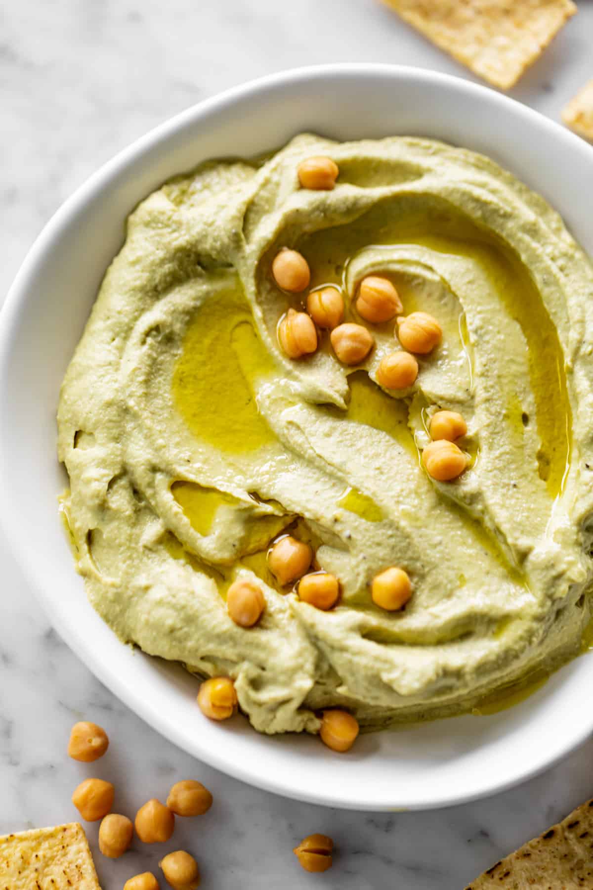 Houmous à l'avocat servi dans un bol blanc, arrosé d'huile d'olive et garni de 16 pois chiches entiers. Des pois chiches entiers décorent le coin inférieur gauche de la table avec des chips de maïs.