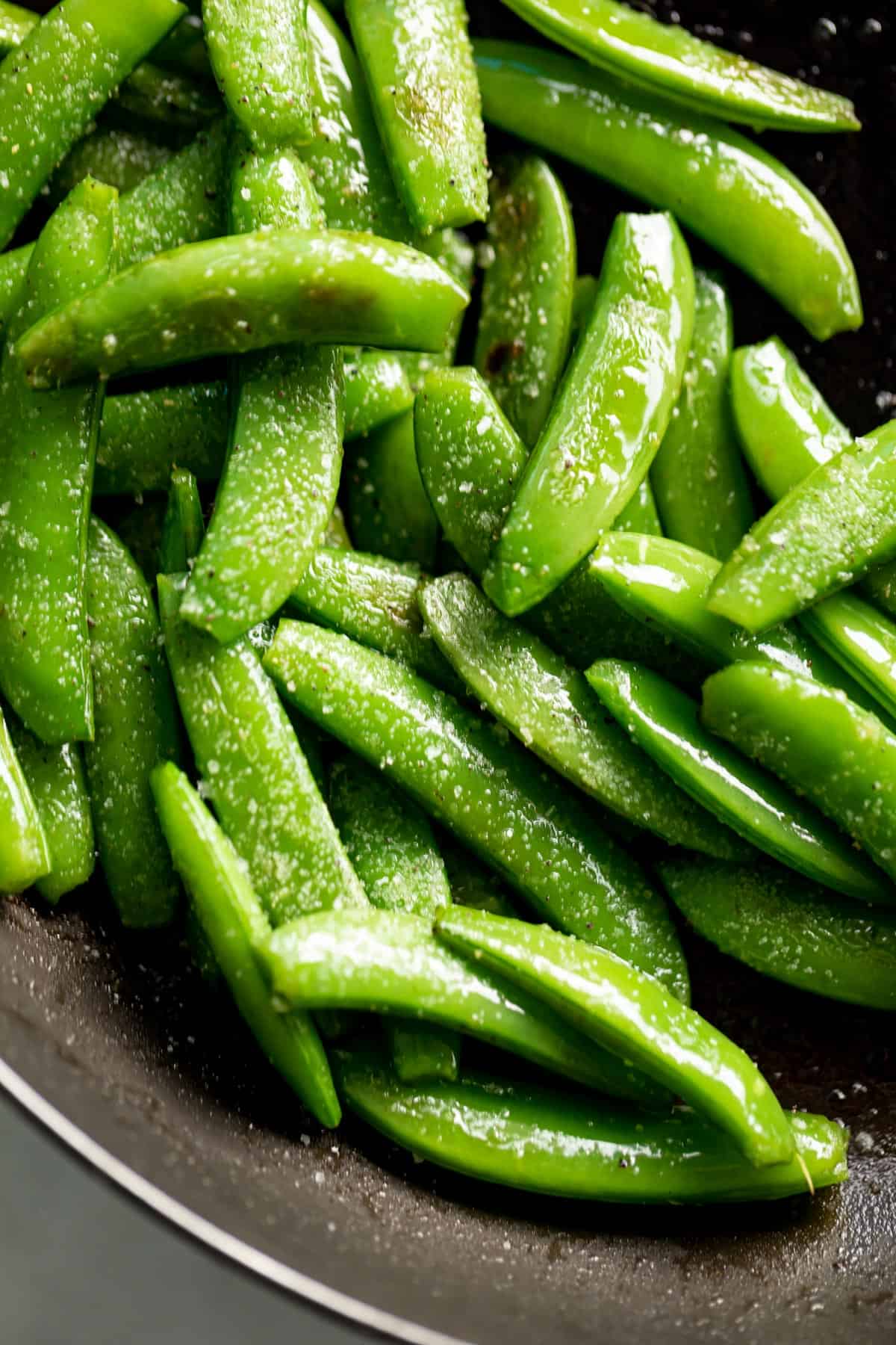 Vue de dessus des pois sucrés sautés dans une poêle noire, saupoudrés de sel gros.