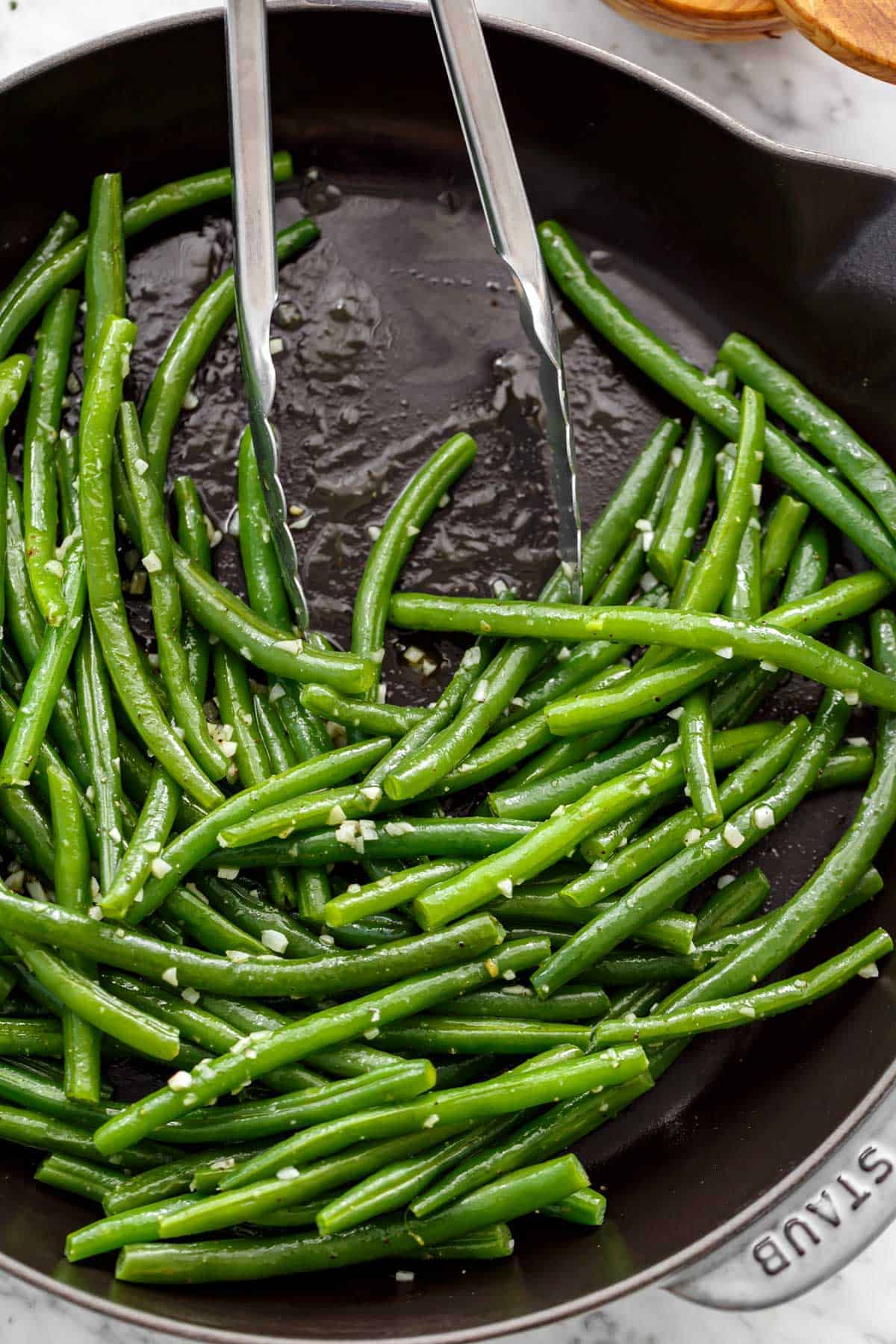 Haricots verts sautés au beurre recouverts d'ail avec des pinces reposant sur le côté de la poêle.