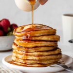 pouring maple syrup on a stack of pancakes.