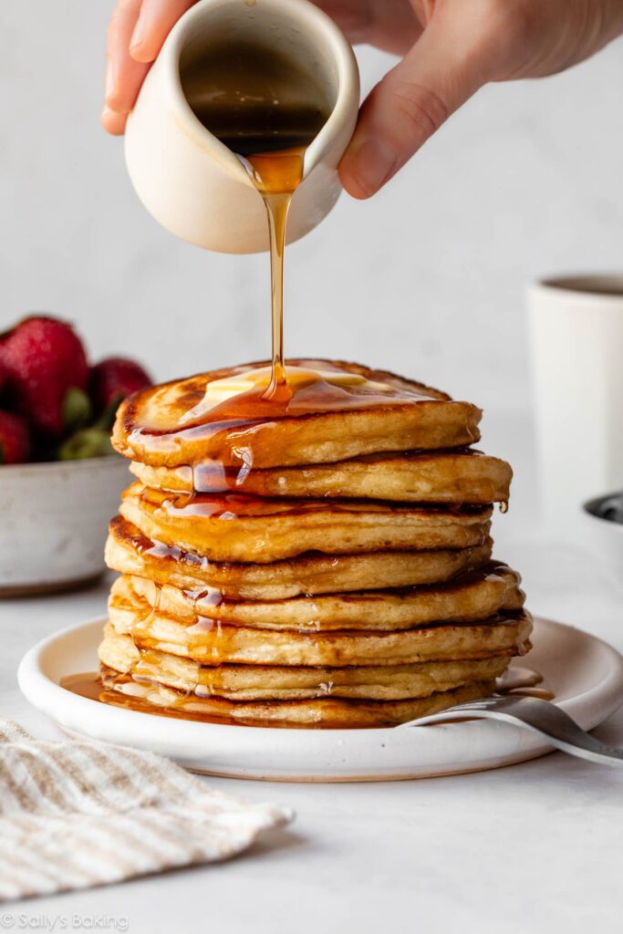 pouring maple syrup on a stack of pancakes.