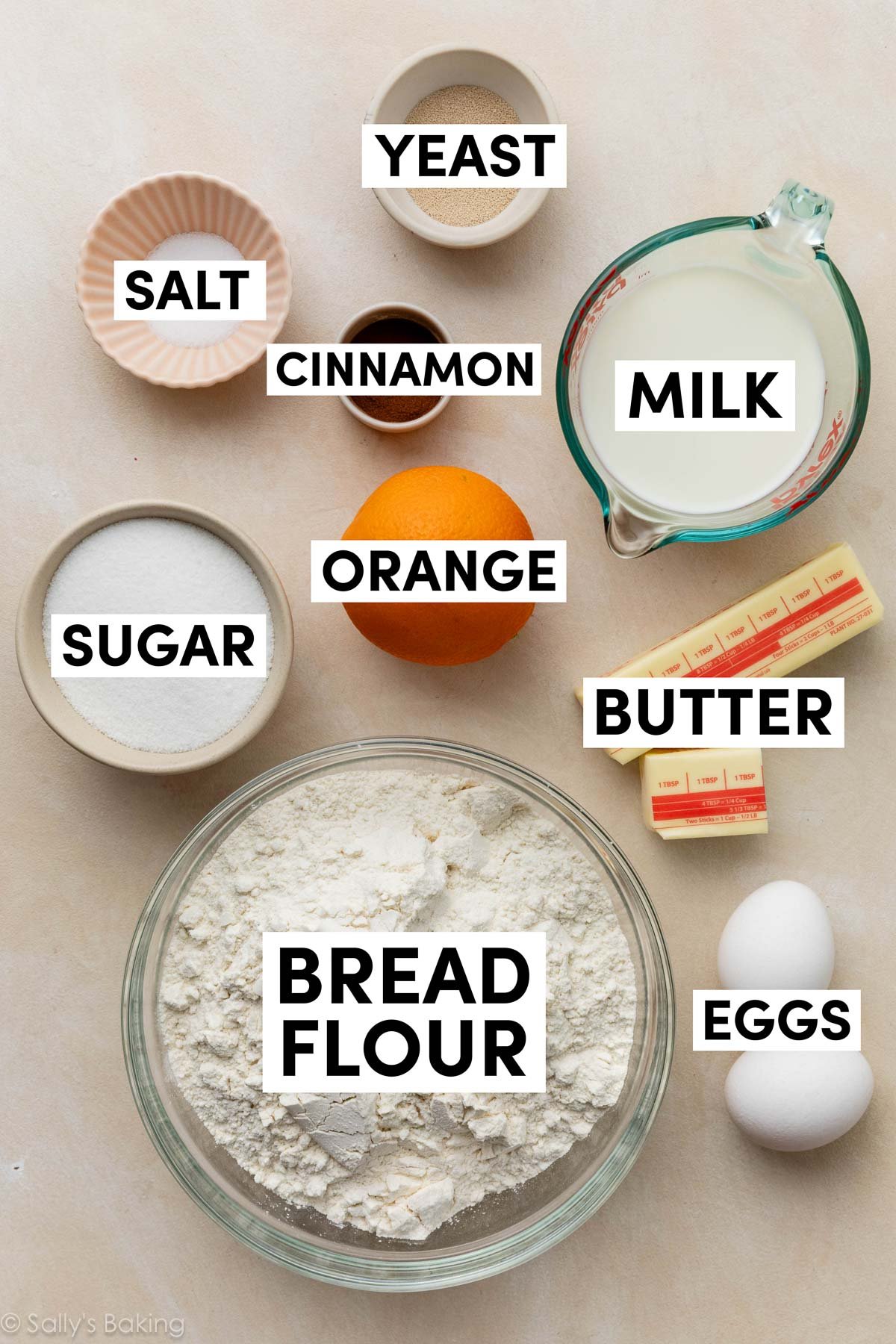 ingrédients sur une surface beige incluant farine à pain, beurre, lait, œufs, cannelle, sucre et levure.