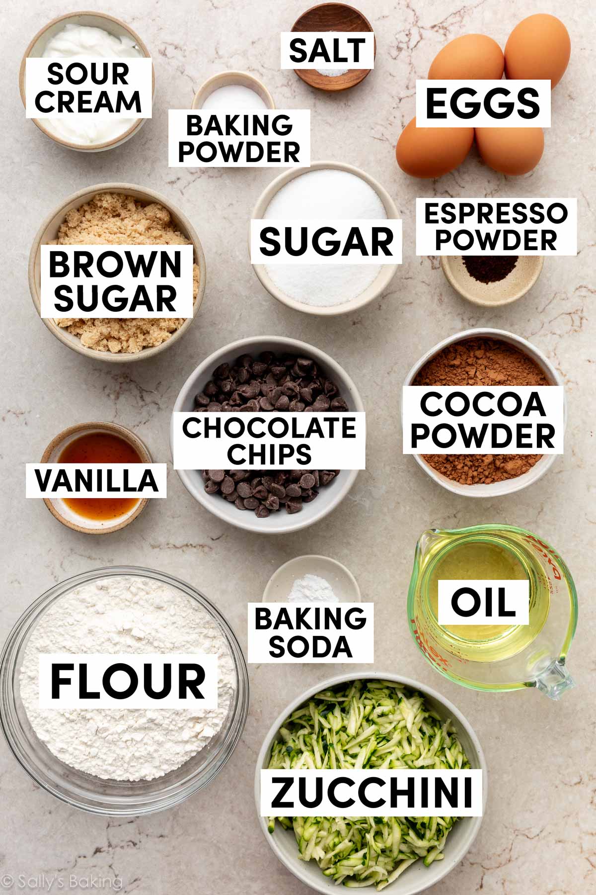 Ingrédients sur une surface marbrée incluant des pépites de chocolat, de la poudre de cacao, de l'huile, de la farine, des œufs, de la poudre d'espresso, du sucre brun, et de la crème aigre.