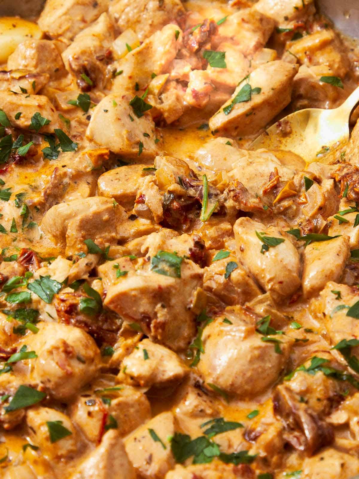 Golden spoon scooping creamy Greek Yogurt Chicken with sun-dried tomatoes and fresh parsley from a rich, velvety sauce.