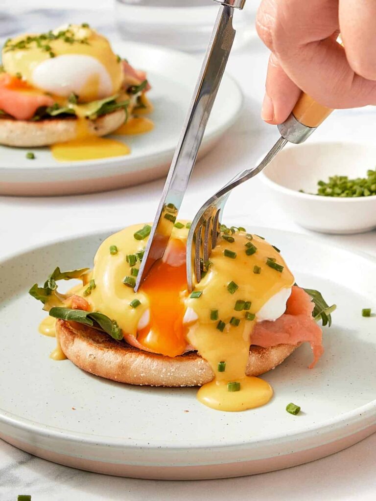 A fork and knife cutting into a poached egg on toasted English muffin with creamy hollandaise sauce and chives — the yolk just starting to spill out.