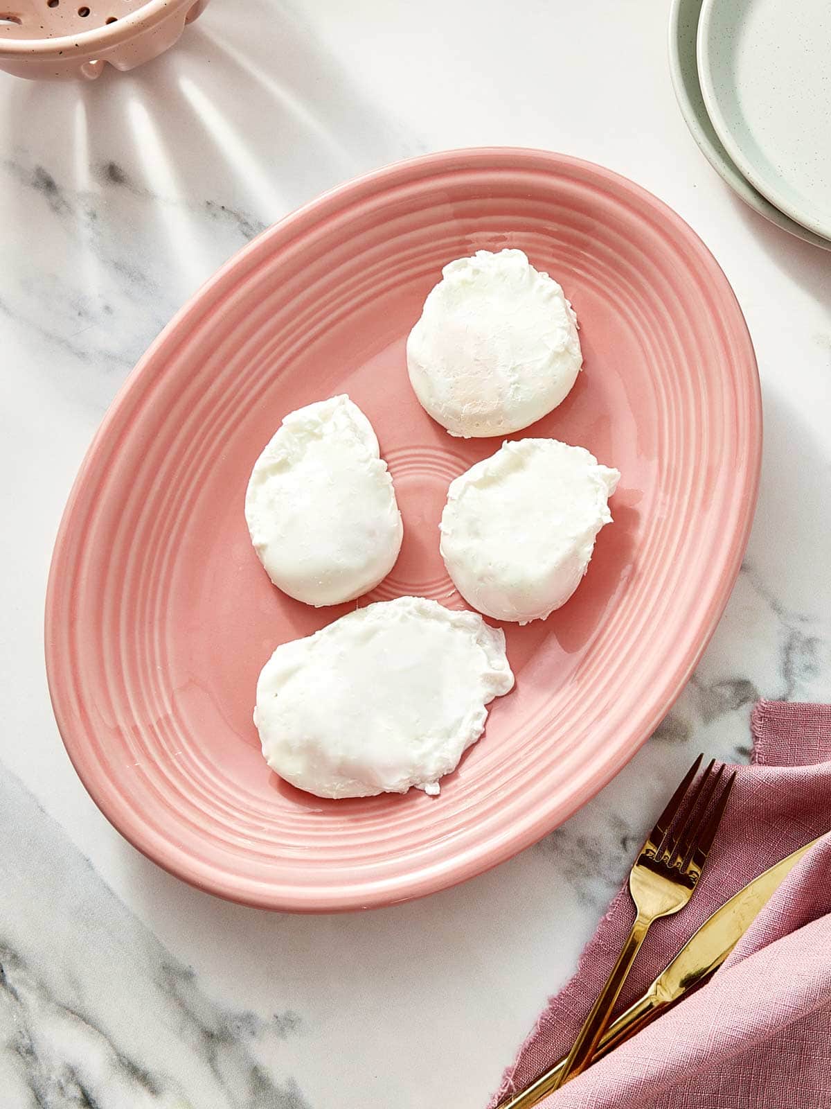 Quatre œufs pochés parfaitement reposant sur une assiette rose pâle sur un fond en marbre.