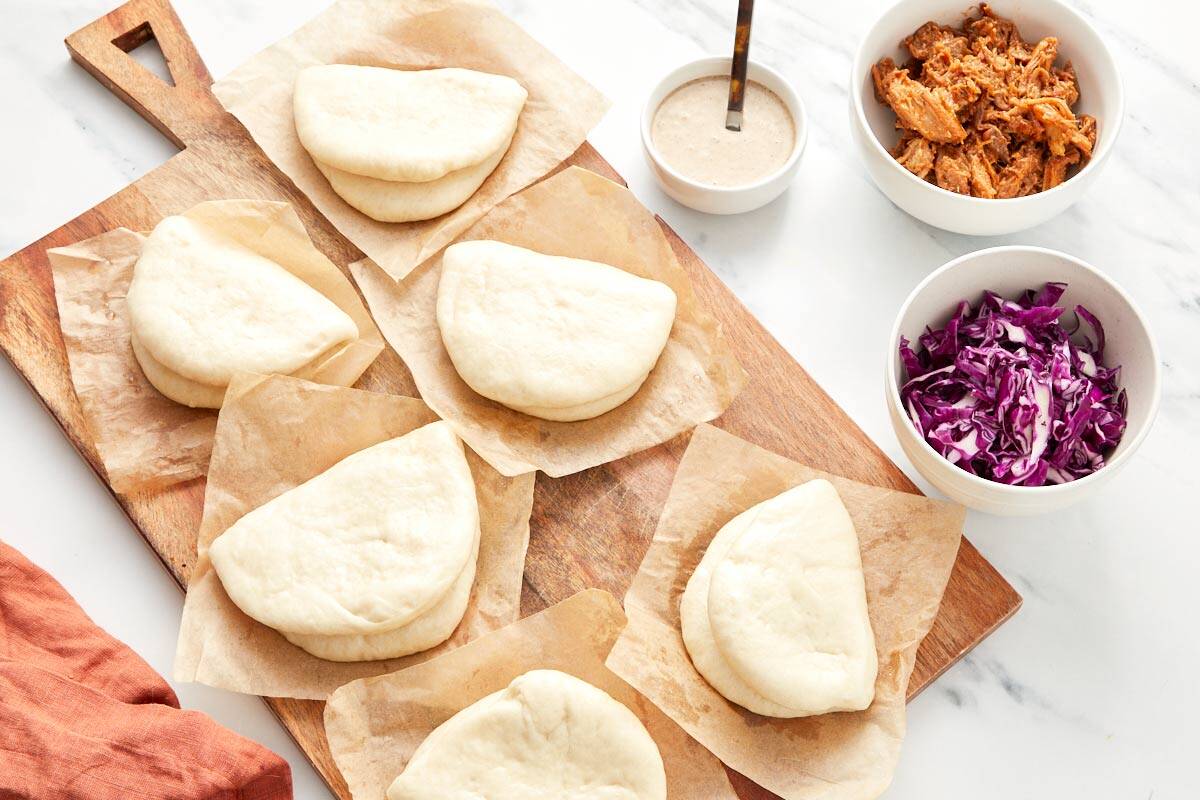 Brioches bao vapeur prêtes à être farcies, à côté de bols de porc effiloché, de chou râpé, et de mayo à l'ail noir.