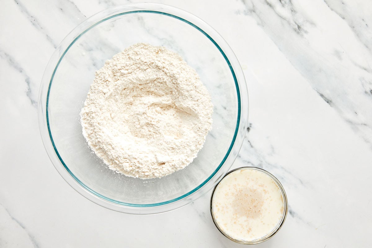 Ingrédients secs pour la pâte à bao dans un bol en verre avec un petit bol de levure mousseuse et de lait tiède à côté.