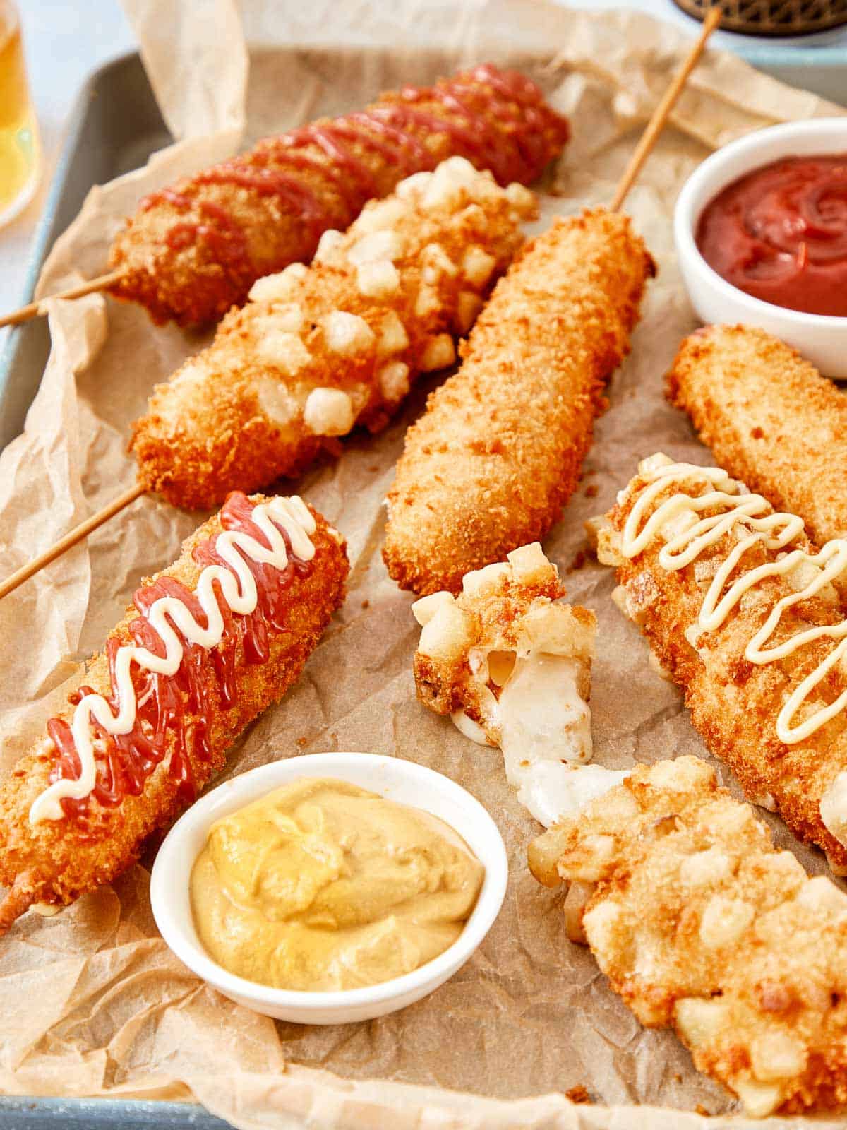 Crispy Korean corn dogs on a tray, drizzled with ketchup and mayo, served with mustard and ketchup dips.