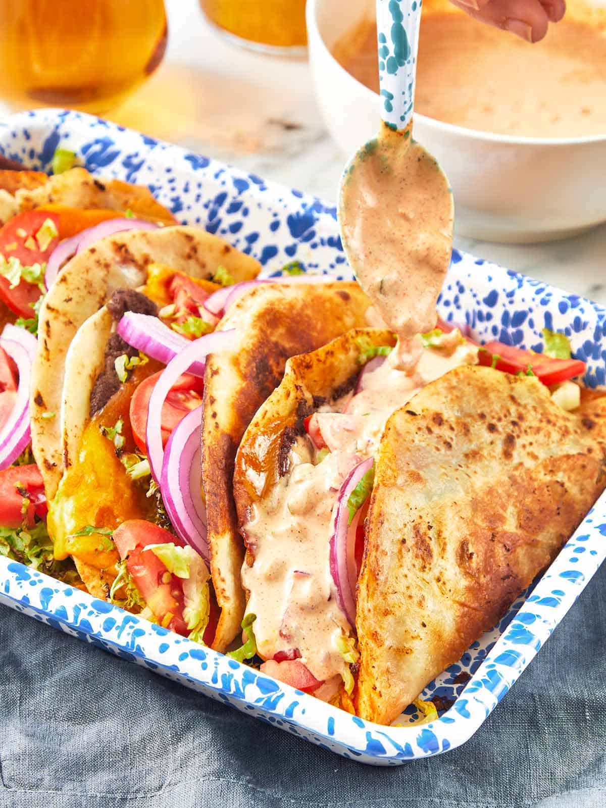 Gros plan sur des tacos de smash burger dans un plateau bleu tacheté, garnis de laitue, tomate, oignon rouge, fromage fondu, et arrosés de sauce crémeuse.