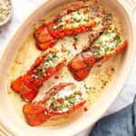 Four broiled lobster tails in a baking dish, topped with chimichurri butter and garnished with fresh herbs and red chili.