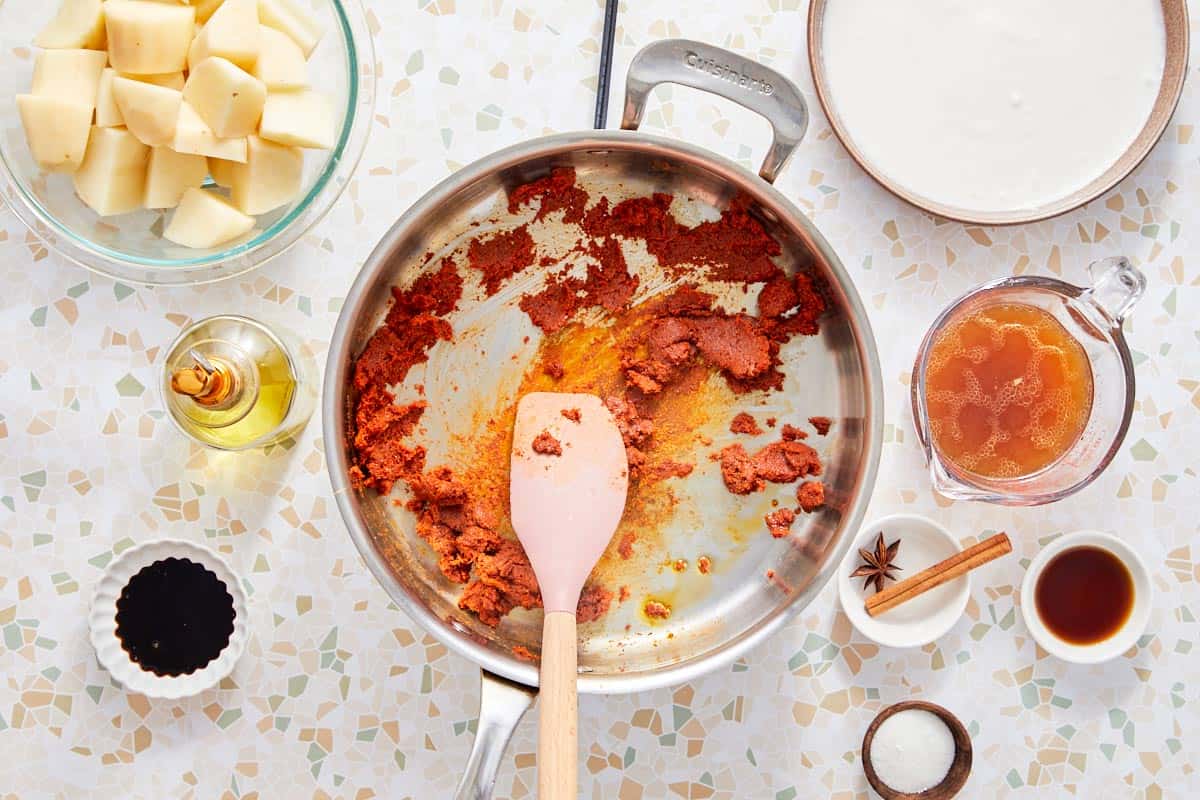 Pâte de curry cuisant dans une poêle avec des pommes de terre, bouillon, et lait de coco à proximité.