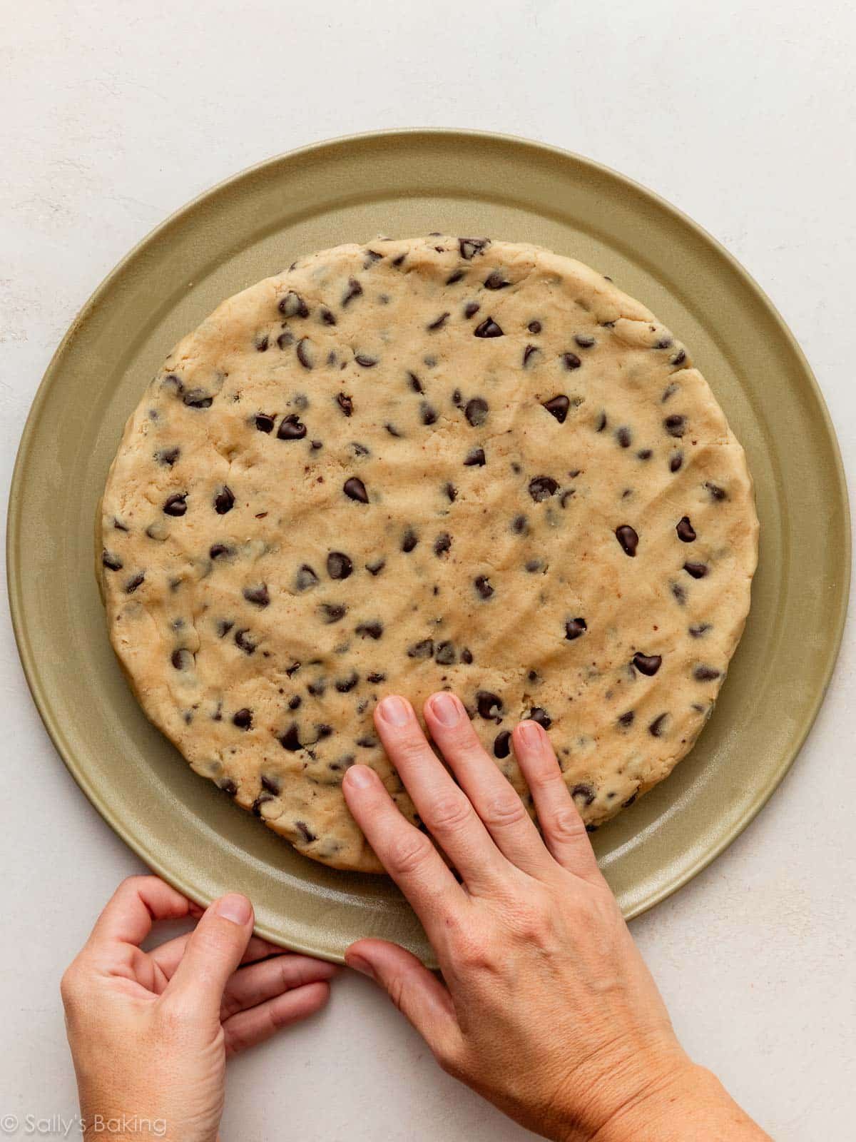 mains aplatissant la pâte à cookies sur une plaque à pizza dorée.