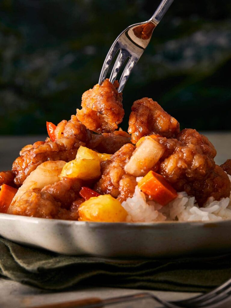 Fork lifting a piece of sweet and sour chicken with pineapple and rice.