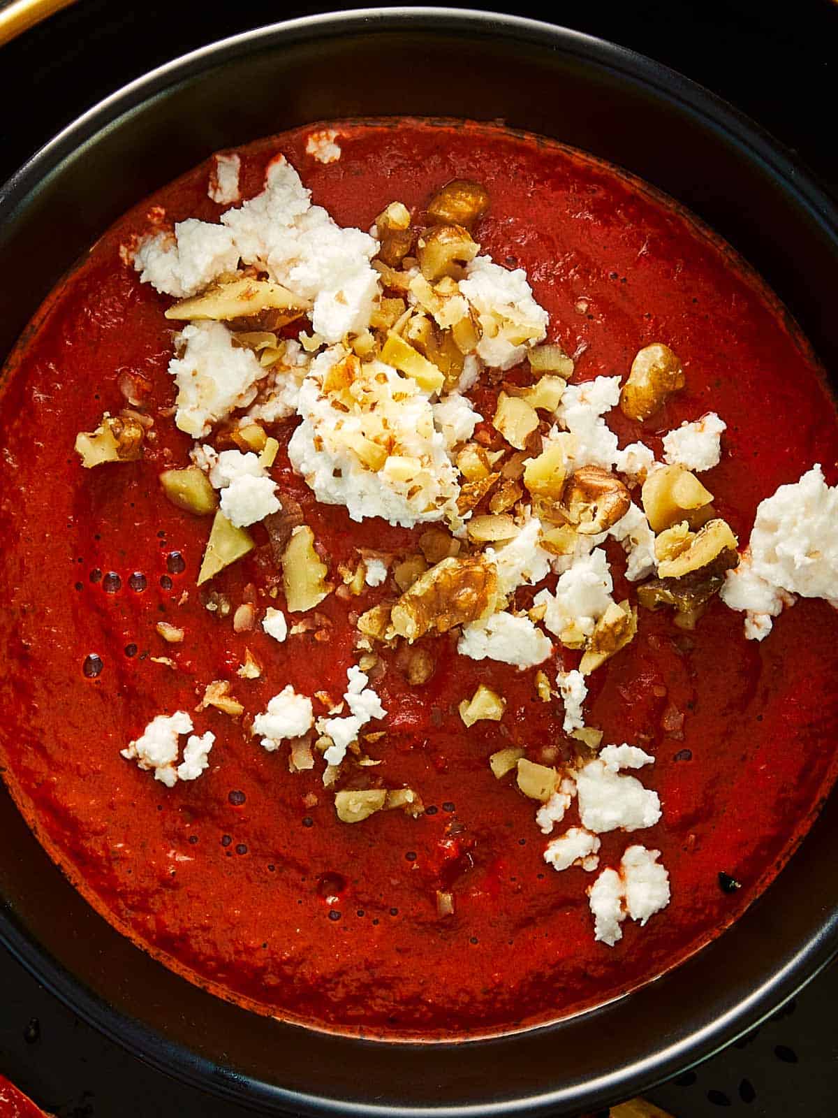 Gros plan d'une trempette rouge riche, garnie de fromage feta émietté et de noix hachées, servie dans un bol noir.