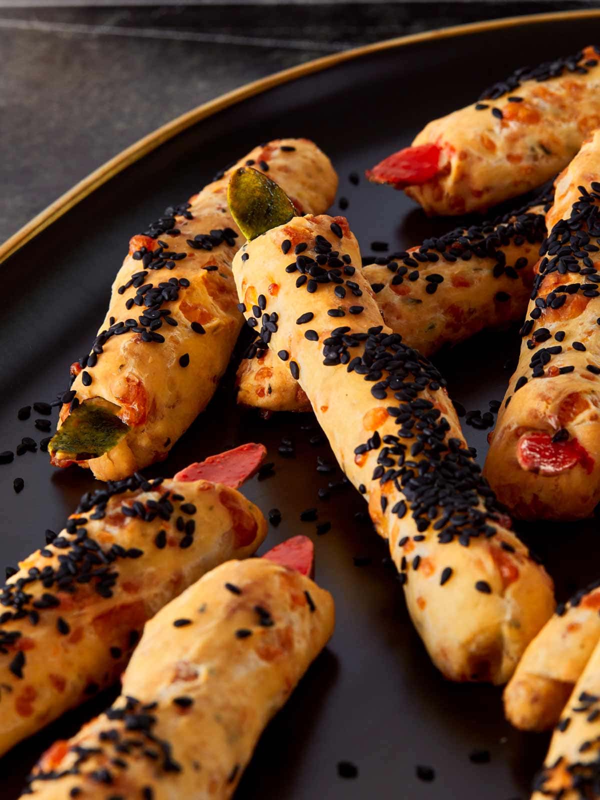 Gros plan sur des Grissini Doigts de Sorcière fraîchement cuits sur une assiette sombre, recouverts de graines de sésame noir et garnis d'amandes rouges et vertes pour un aspect effrayant de Halloween.