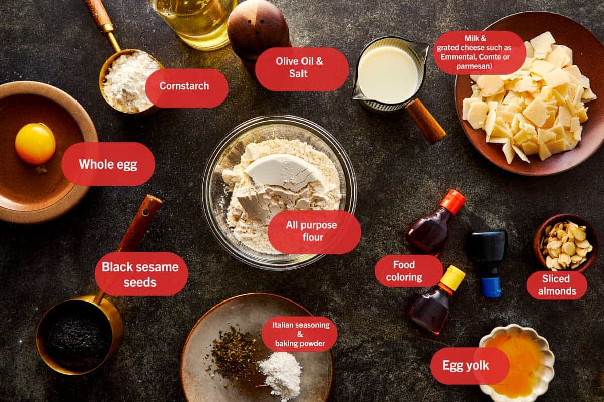Plan aérien des ingrédients des Grissini Doigts de Sorcière disposés sur une surface foncée, y compris de la farine, de la fécule de maïs, du fromage râpé, de l'huile d'olive, du lait, un œuf, des graines de sésame noir, des amandes tranchées, des herbes italiennes et des colorants alimentaires.