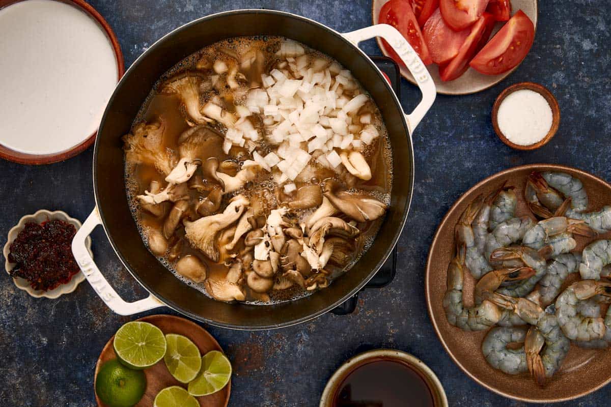 Pot avec des oignons et des champignons d'huître mijotant dans le bouillon, entouré de crevettes, de tomates, de citrons verts et de lait de coco.