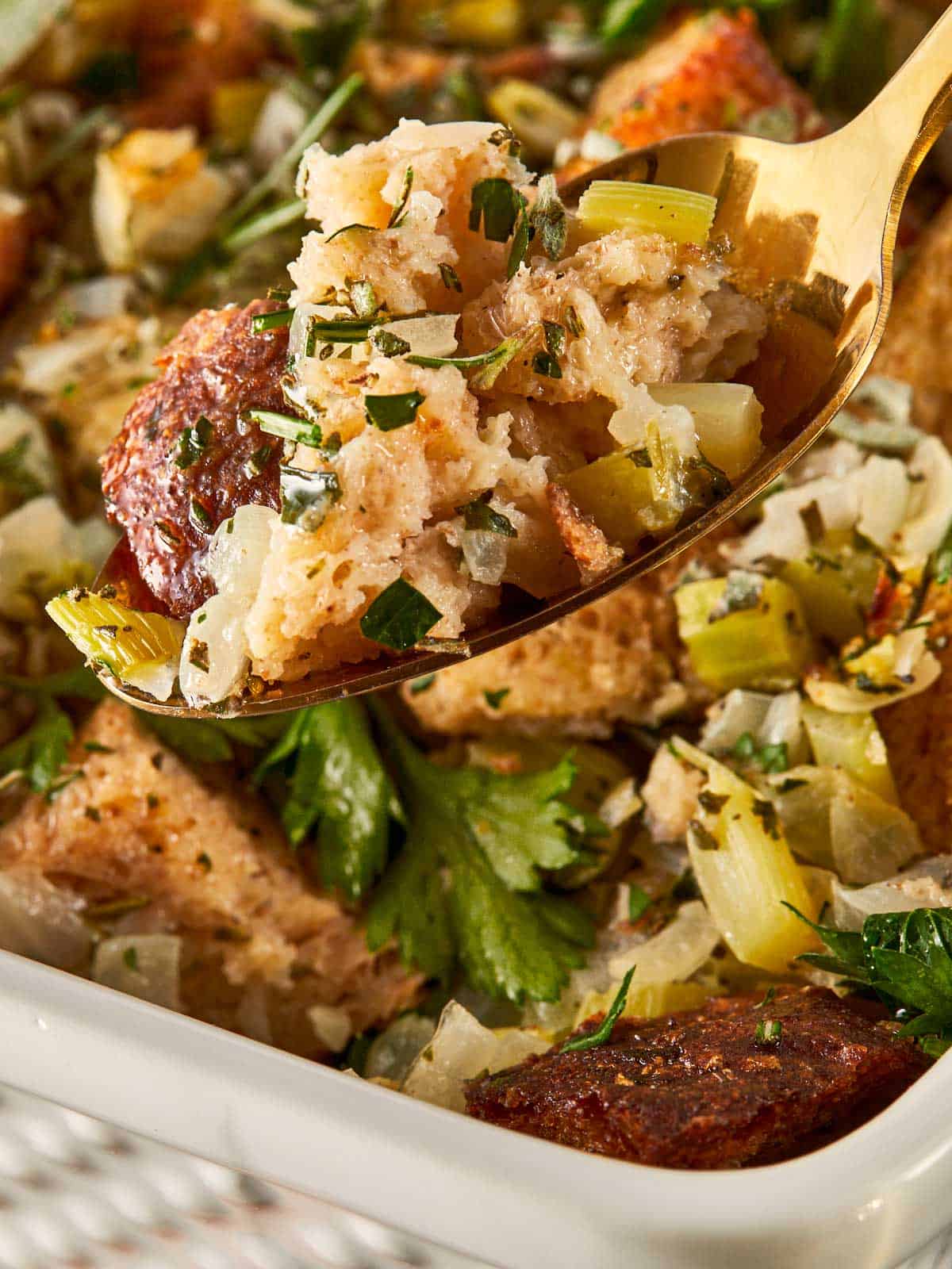 Cubes dorés de farce beurrée aux herbes dans un plat de cuisson blanc, surmontés de persil frais, de sauge et de romarin. Une cuillère dorée soulève une bouchée parfaite montrant le centre moelleux et les bords croustillants.