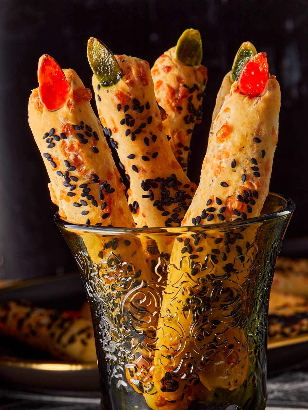 Gros plan sur des grissini en forme de doigts de sorcière dans un verre décoratif, garnis d'amandes rouges et vertes et recouverts de graines de sésame noir pour un aspect effrayant de Halloween.