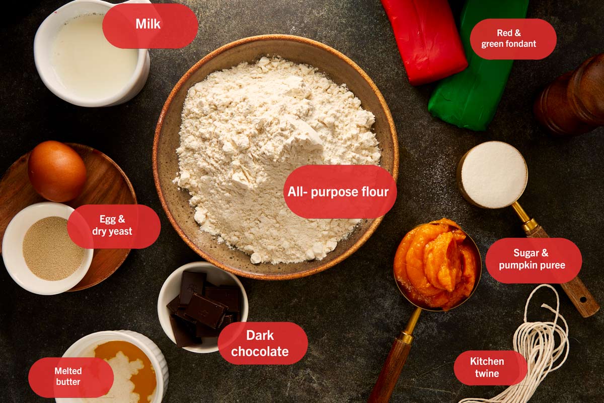 Ingrédients pour les pains à la citrouille disposés sur une surface sombre, comprenant farine, purée de citrouille, lait, chocolat, beurre, œuf, levure, sucre, et fondant.