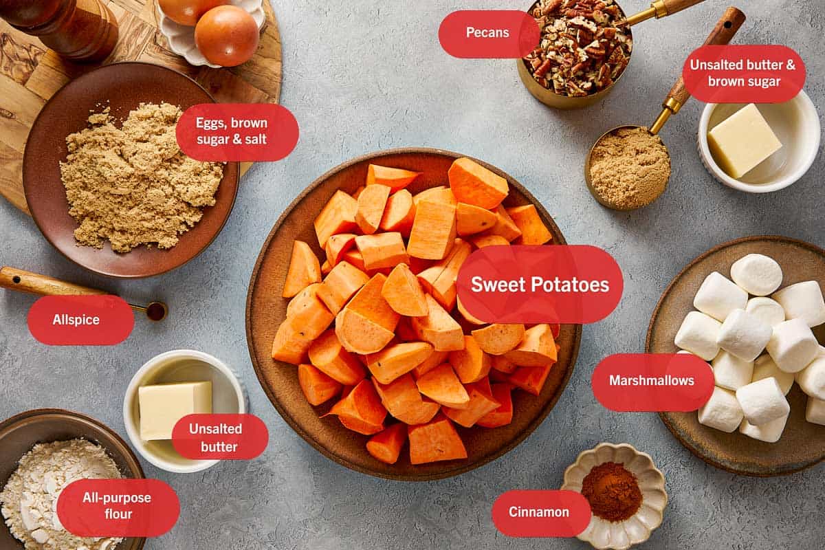 Ingrédients pour la casserole de patates douces disposés, y compris des patates douces épluchées, des guimauves, des noix de pécan, du sucre brun, du beurre, des œufs, de la farine, de la cannelle et du piment de la Jamaïque.